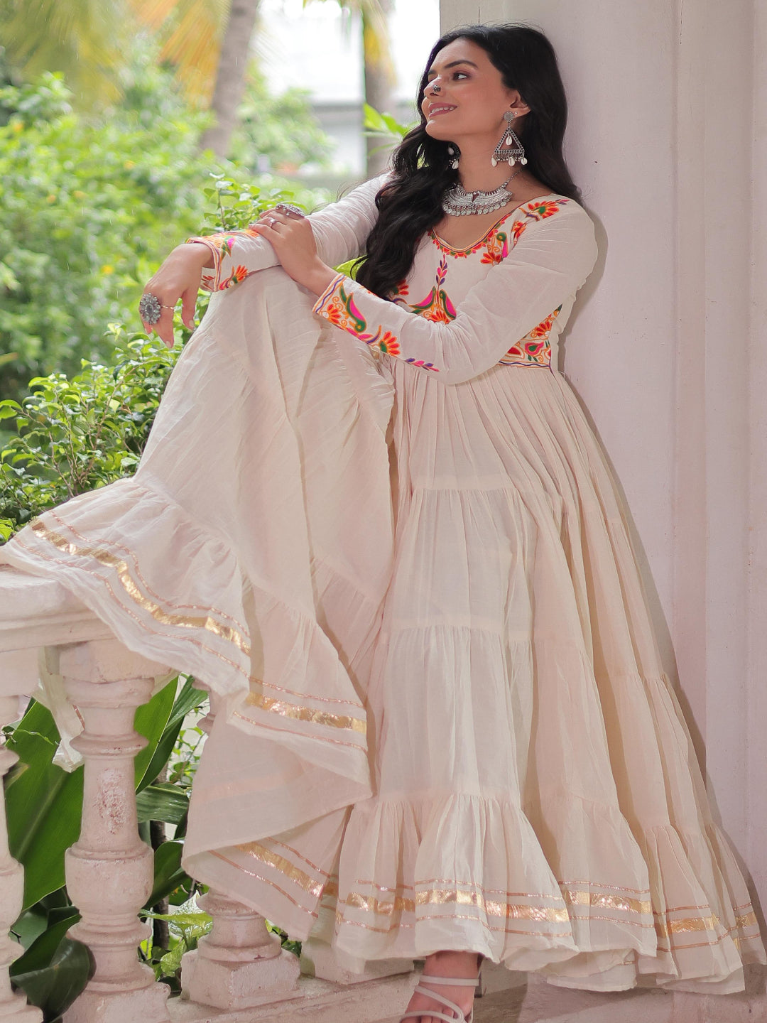 Woman in a white traditional outfit with floral embroidery, standing outdoors with greenery in the background.