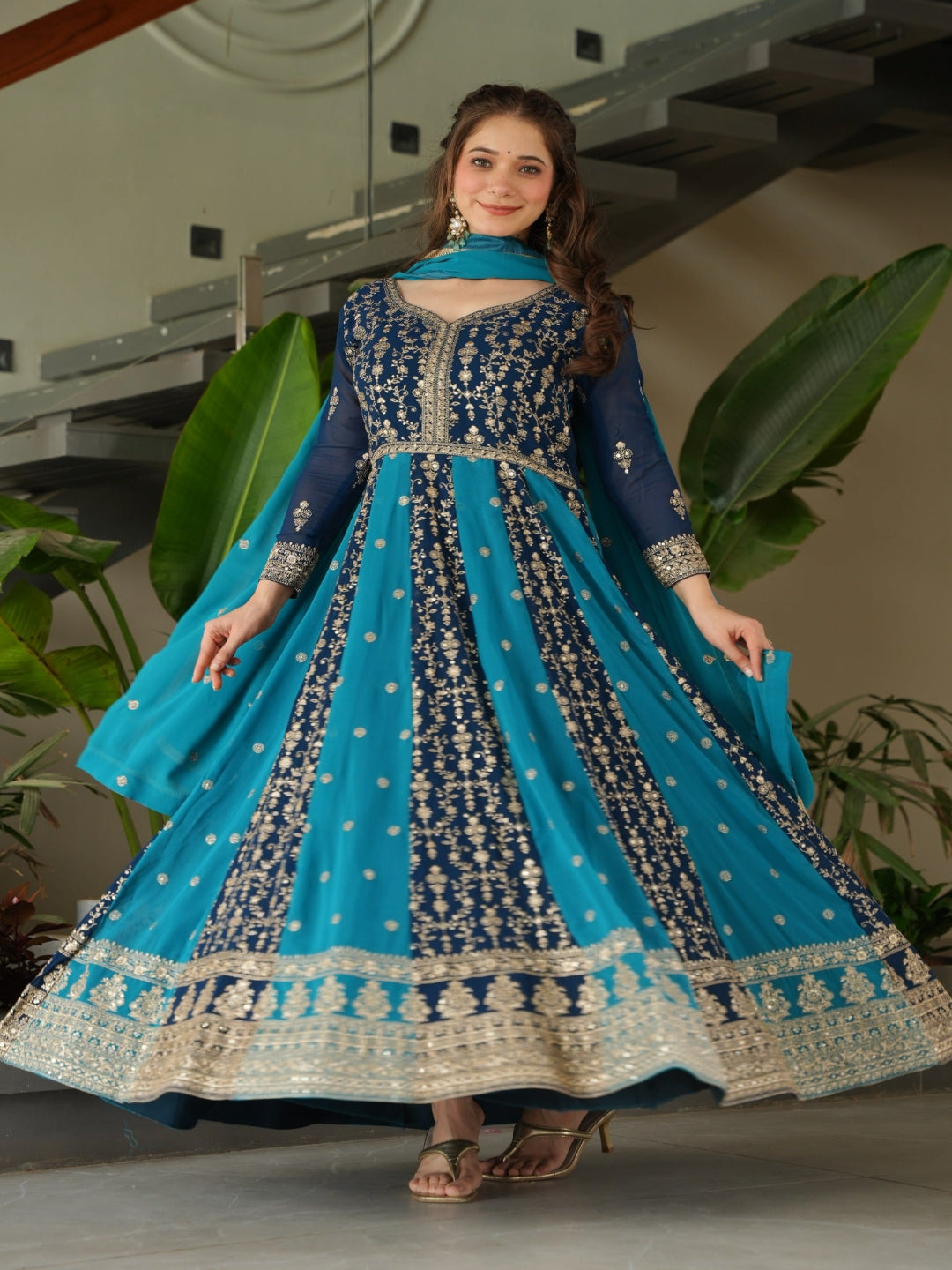 Woman in a blue and black embroidered traditional outfit standing indoors with plants in the background