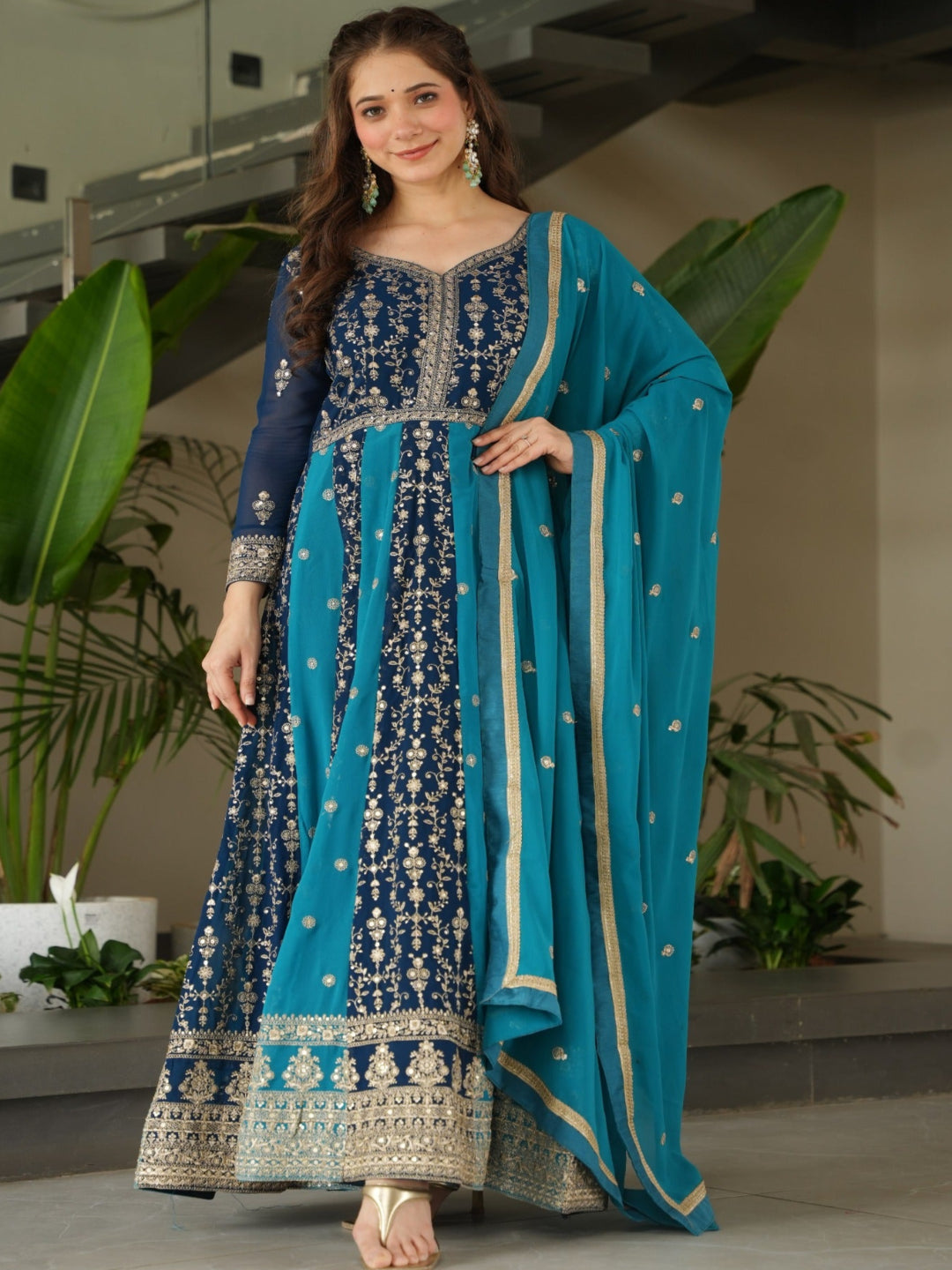 Woman in a blue and navy traditional outfit standing indoors with plants in the background