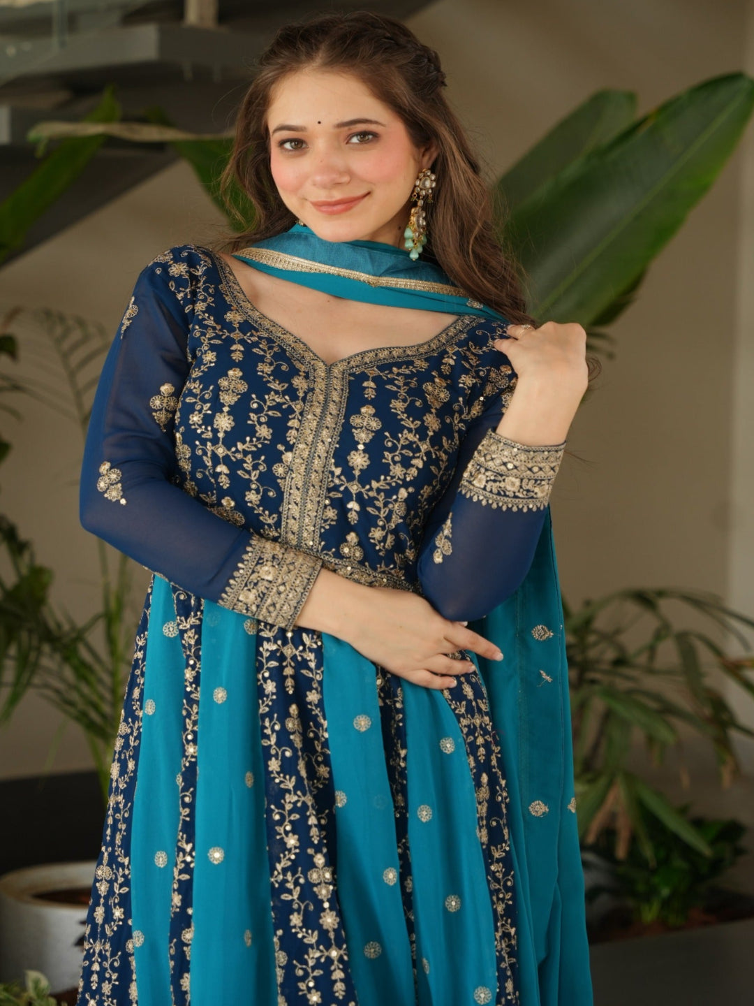 Woman in a blue and silver traditional outfit standing indoors with plants in the background