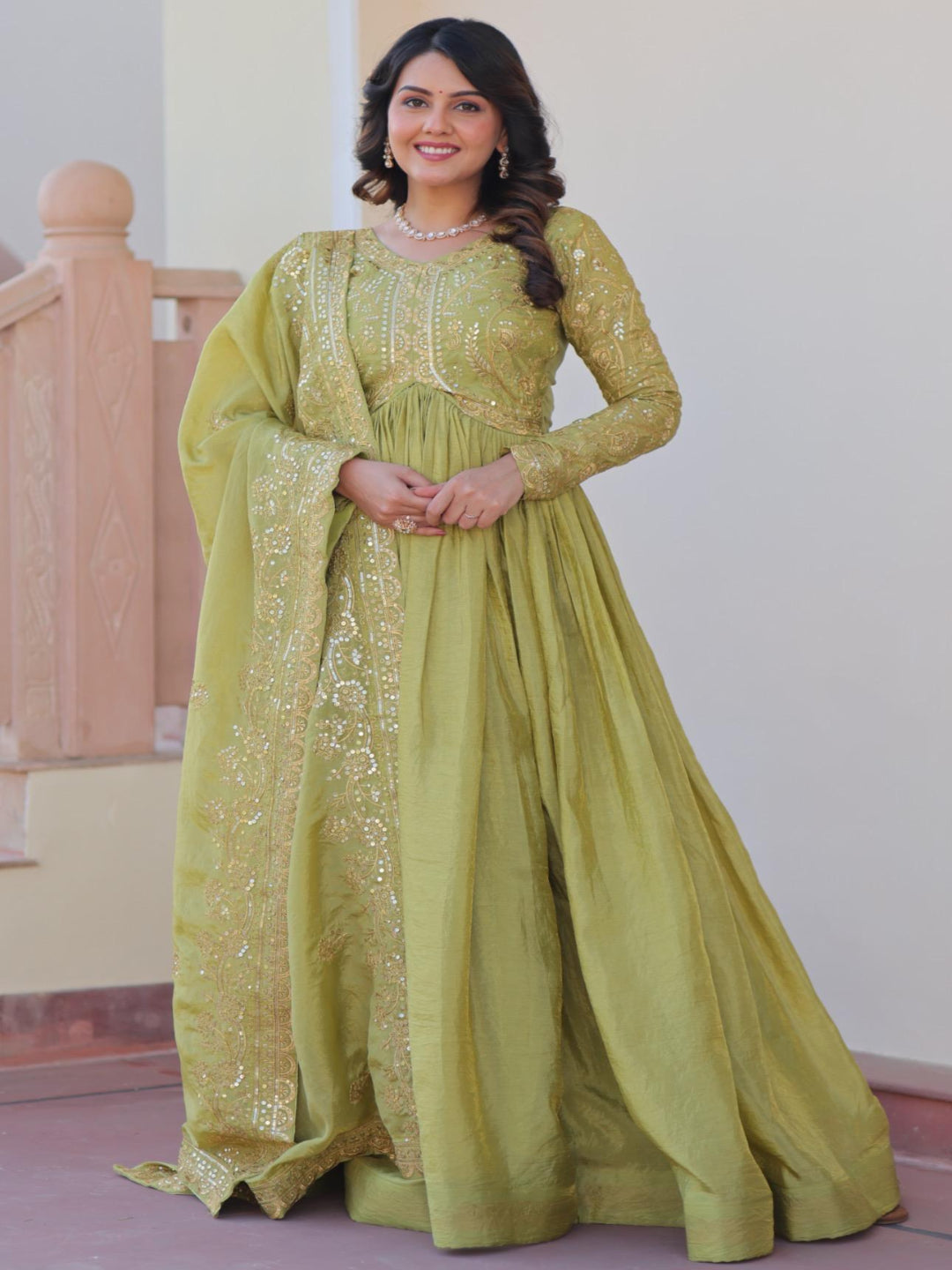 Woman in a green traditional outfit standing indoors with a staircase in the background