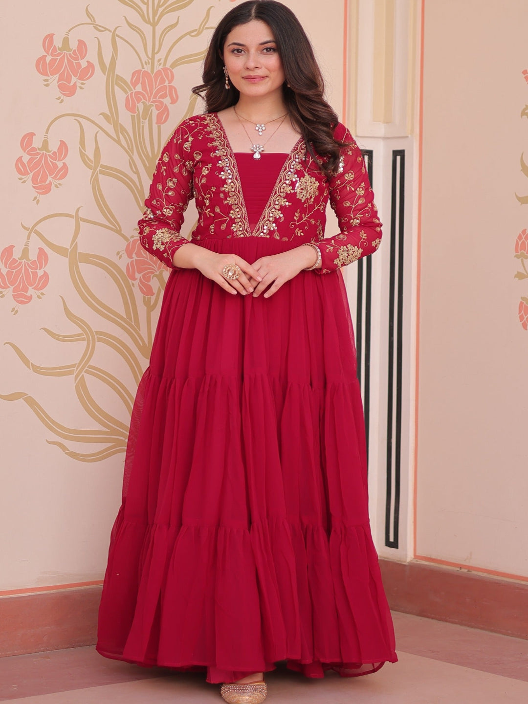 Woman wearing a red traditional outfit with intricate designs against a decorative wall.