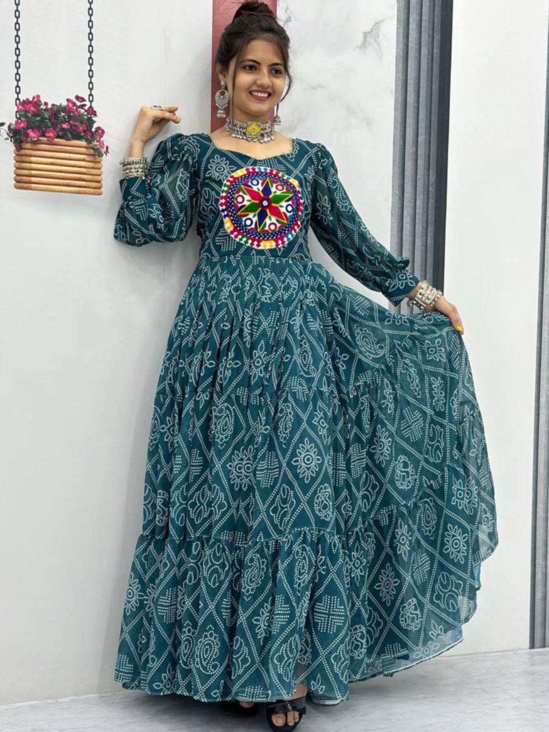 Woman wearing a long teal dress with a colorful floral design, standing indoors.