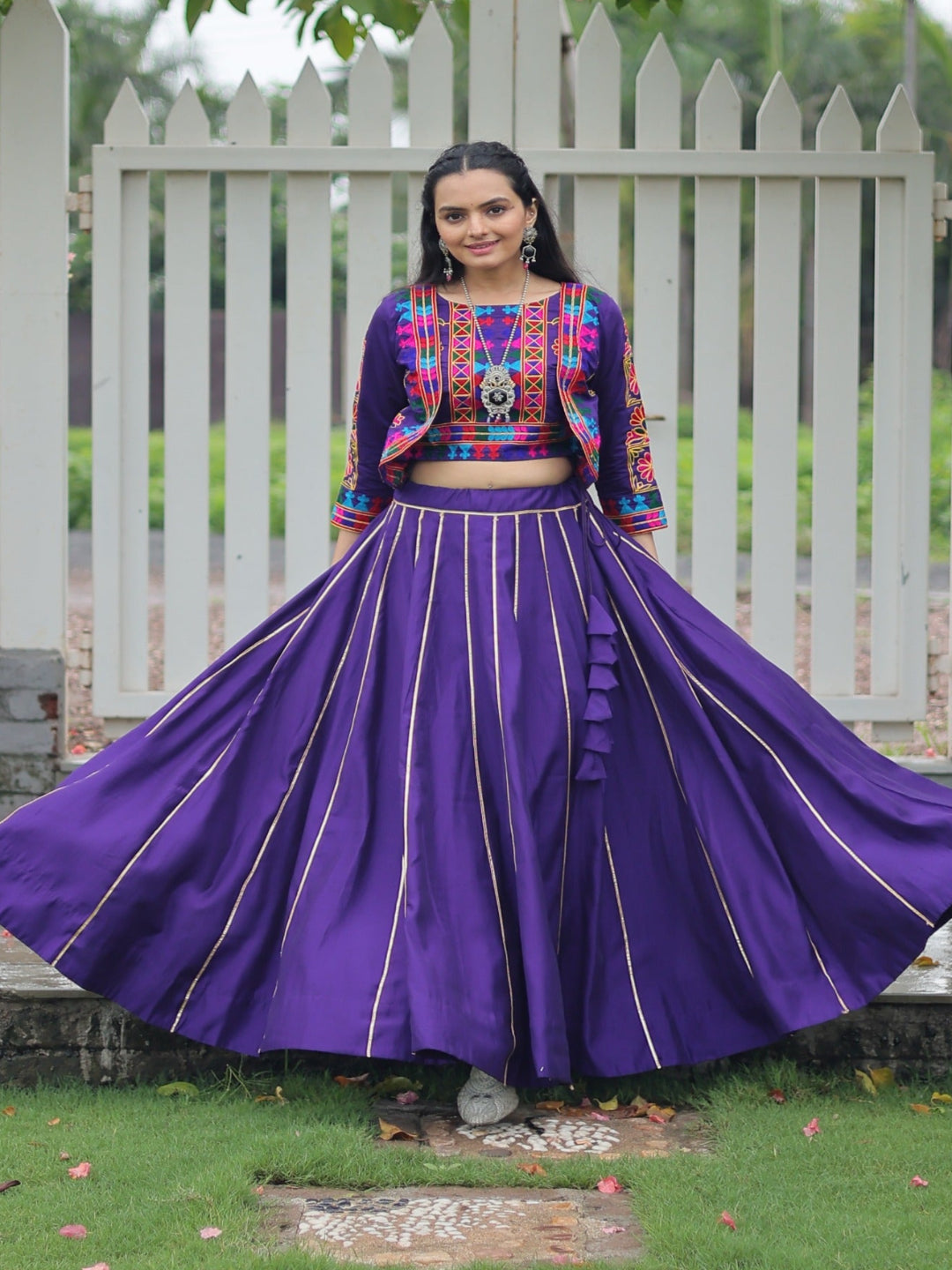 Woman in a colorful traditional outfit with a purple skirt standing outdoors.