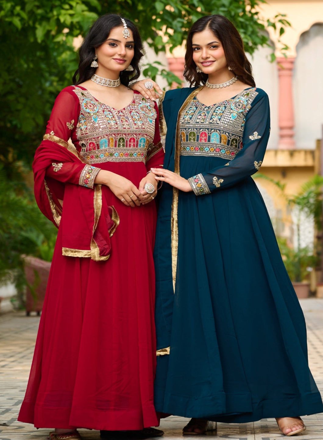 Two women in traditional outfits standing outdoors with greenery in the background