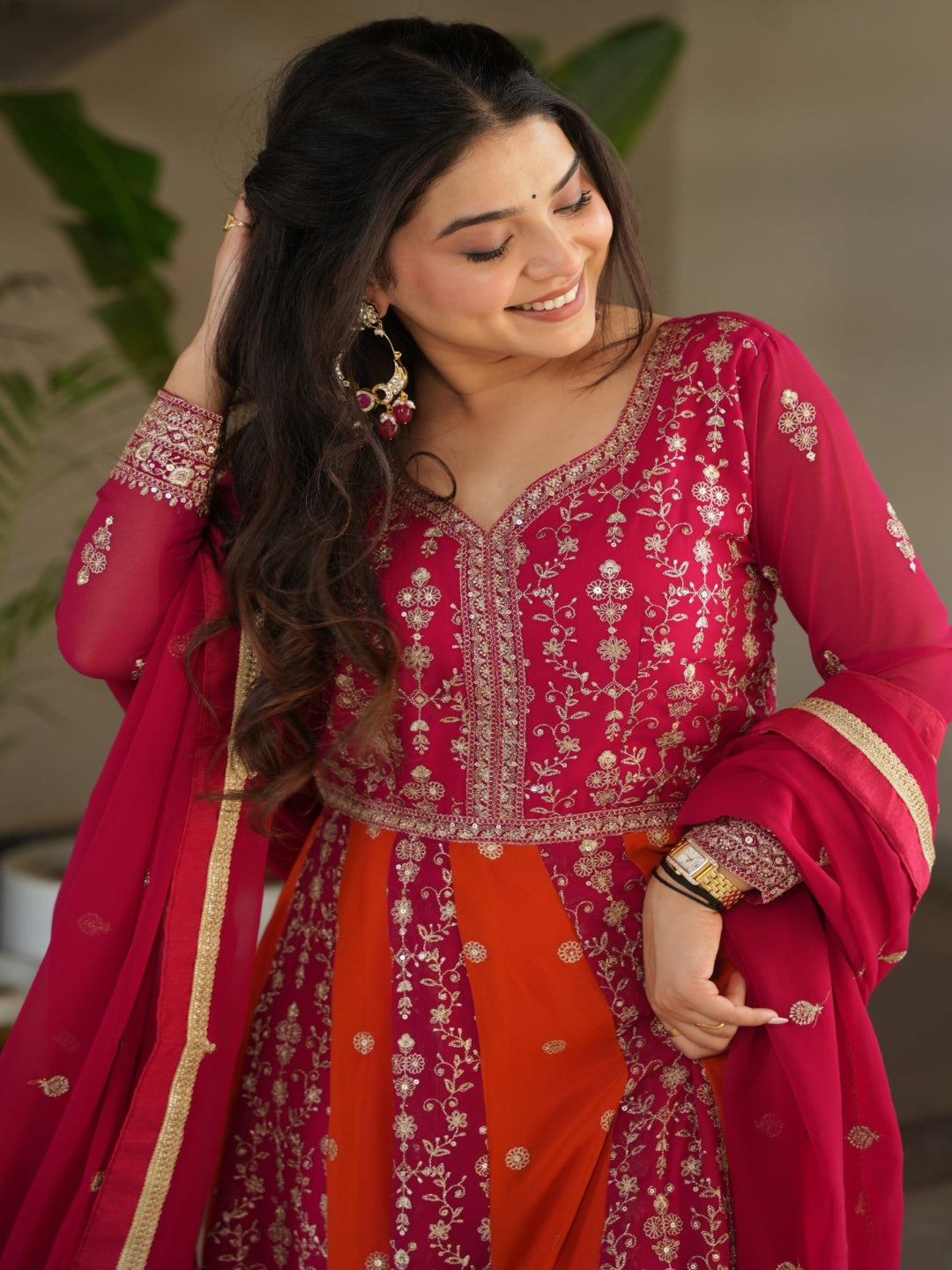 Woman in a traditional pink and red outfit with intricate designs, standing indoors.