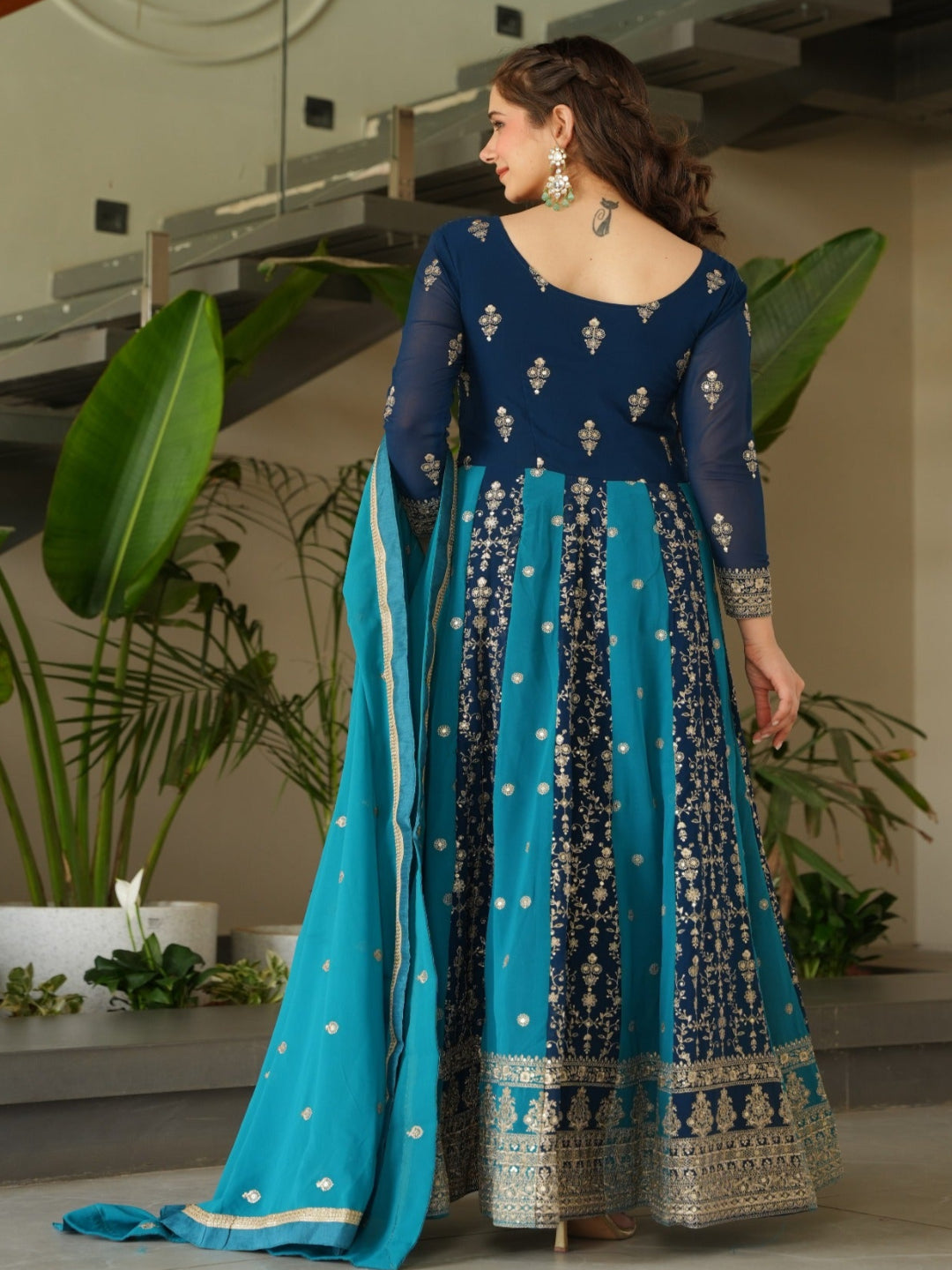Woman in a blue and green traditional outfit standing in a modern indoor setting with plants.