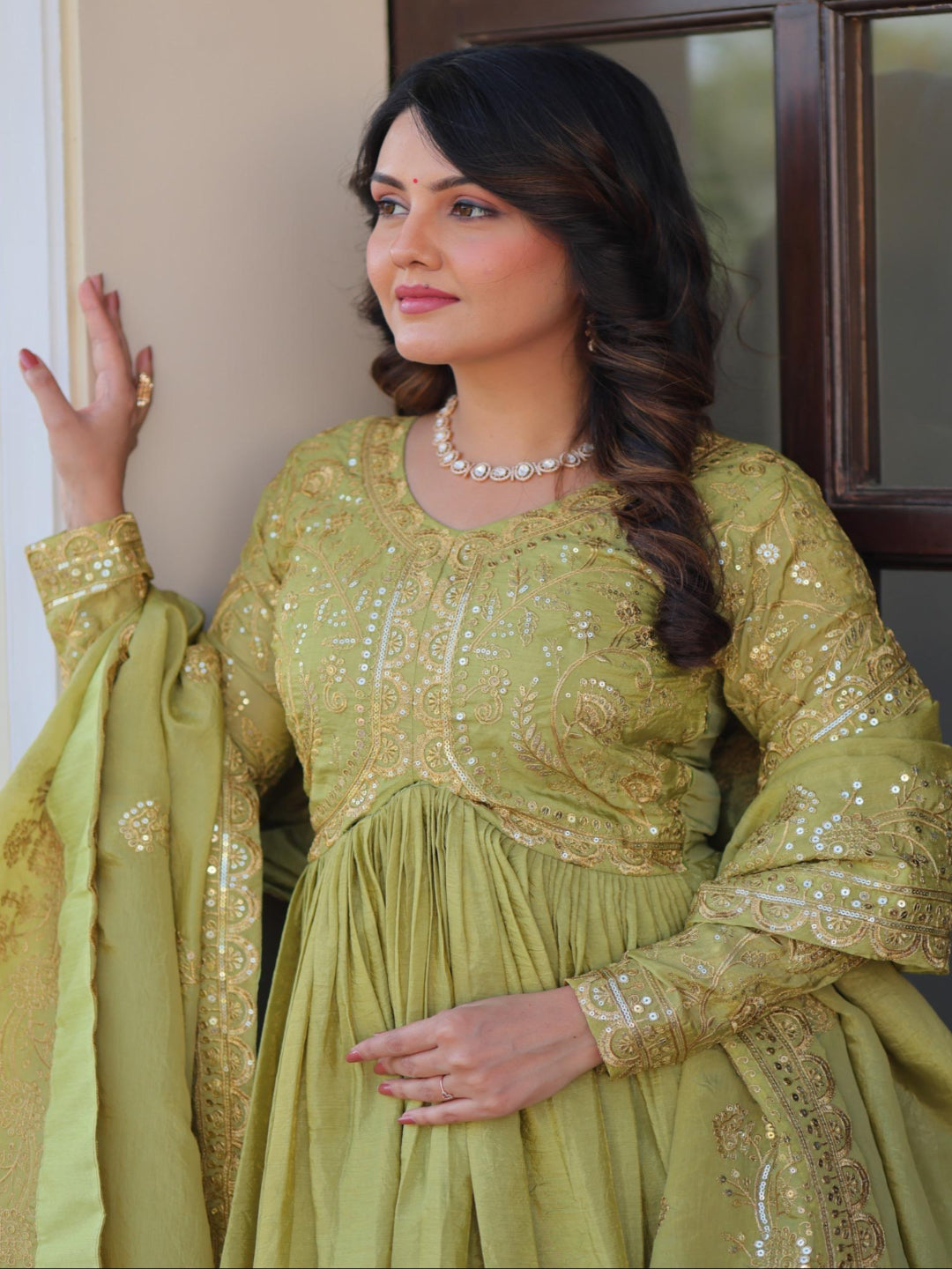Woman in a green traditional outfit with gold embroidery standing indoors.