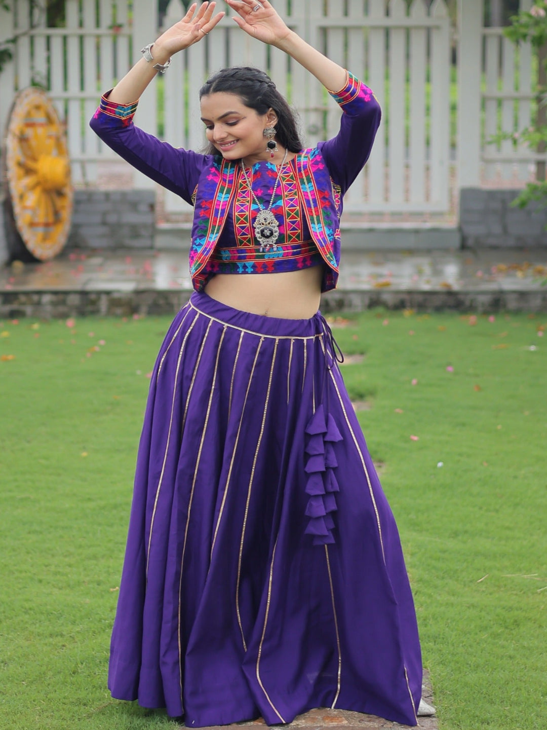 Woman in traditional outfit with colorful top and purple skirt standing in a garden.