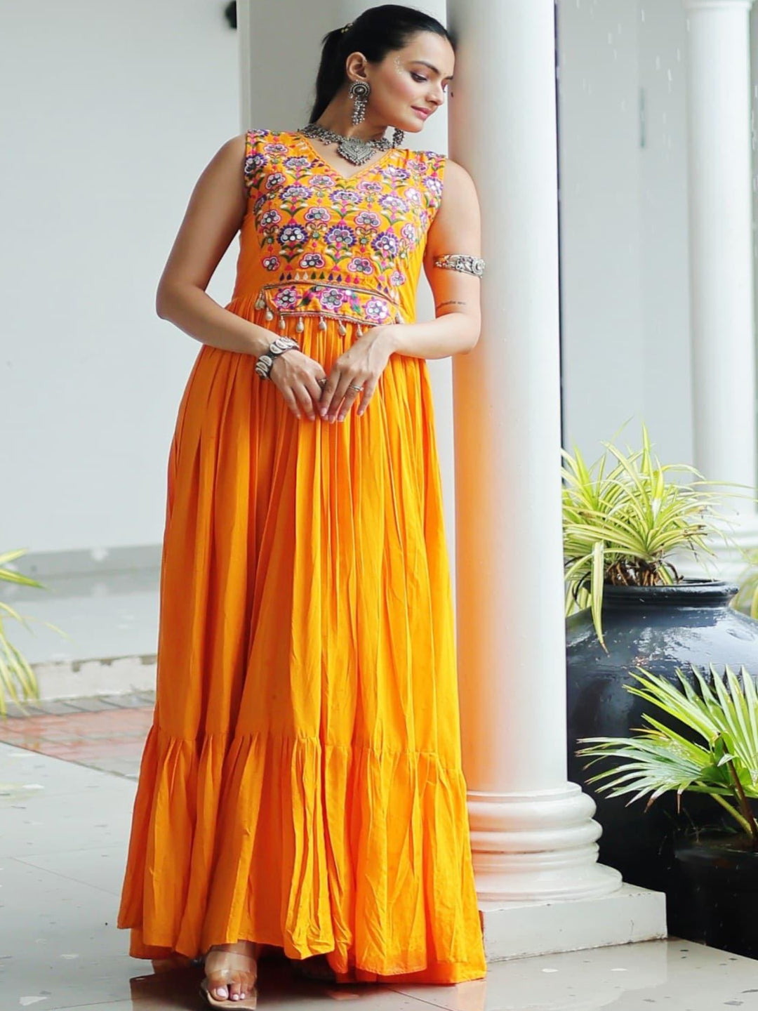 Woman in an orange and patterned dress standing in a white architectural setting with columns and plants.