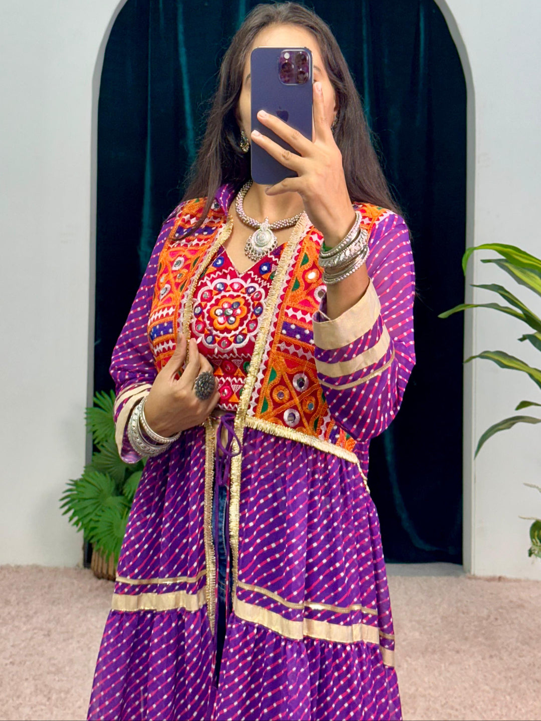 Person wearing a colorful traditional outfit taking a mirror selfie with a plant in the background.