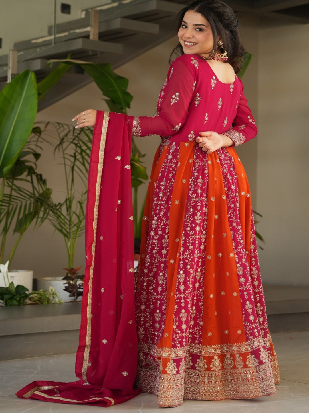 Woman in a red and orange traditional outfit standing indoors with plants and a staircase in the background.