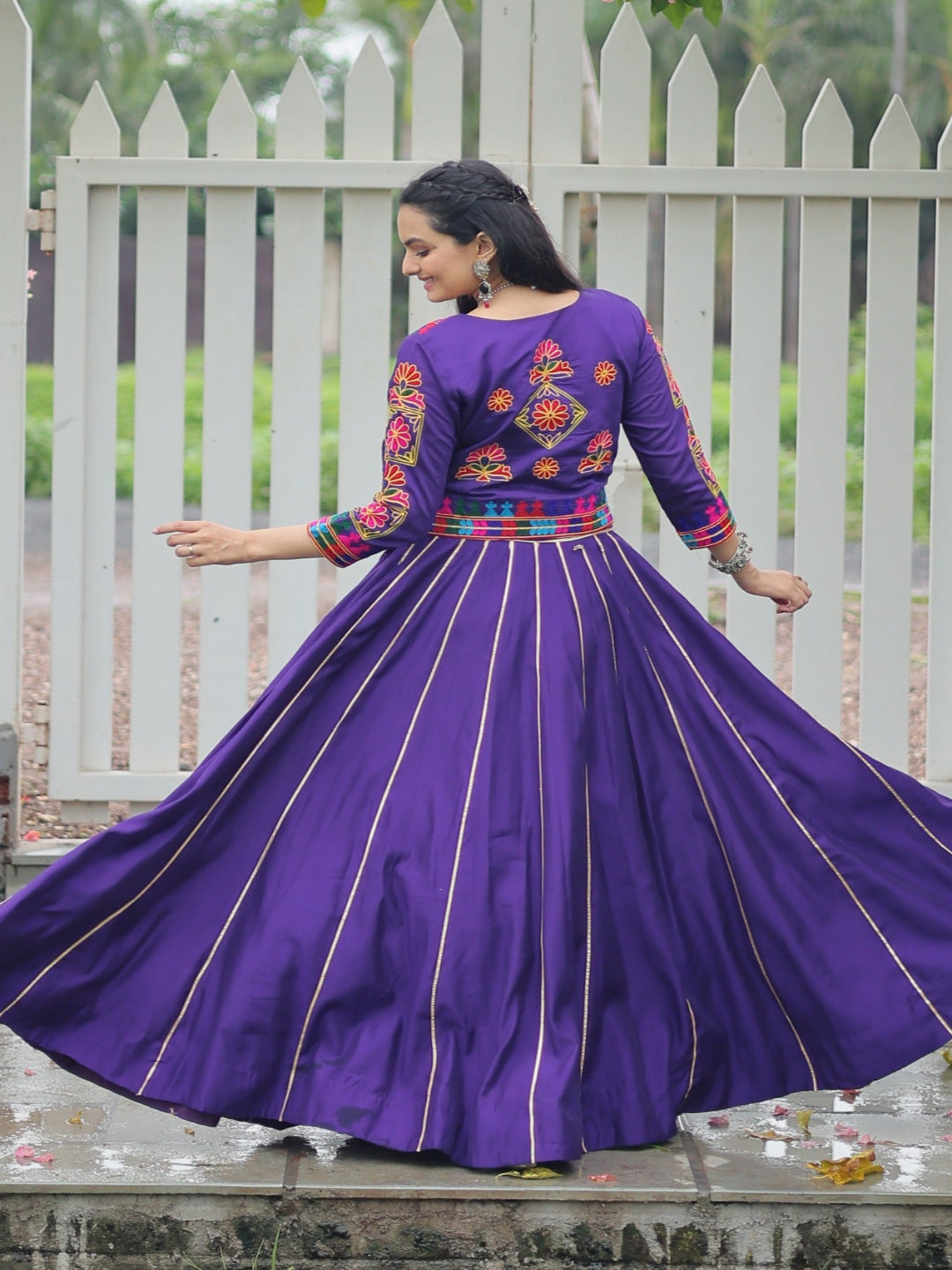 Woman in a purple embroidered dress standing in front of a white picket fence with greenery.