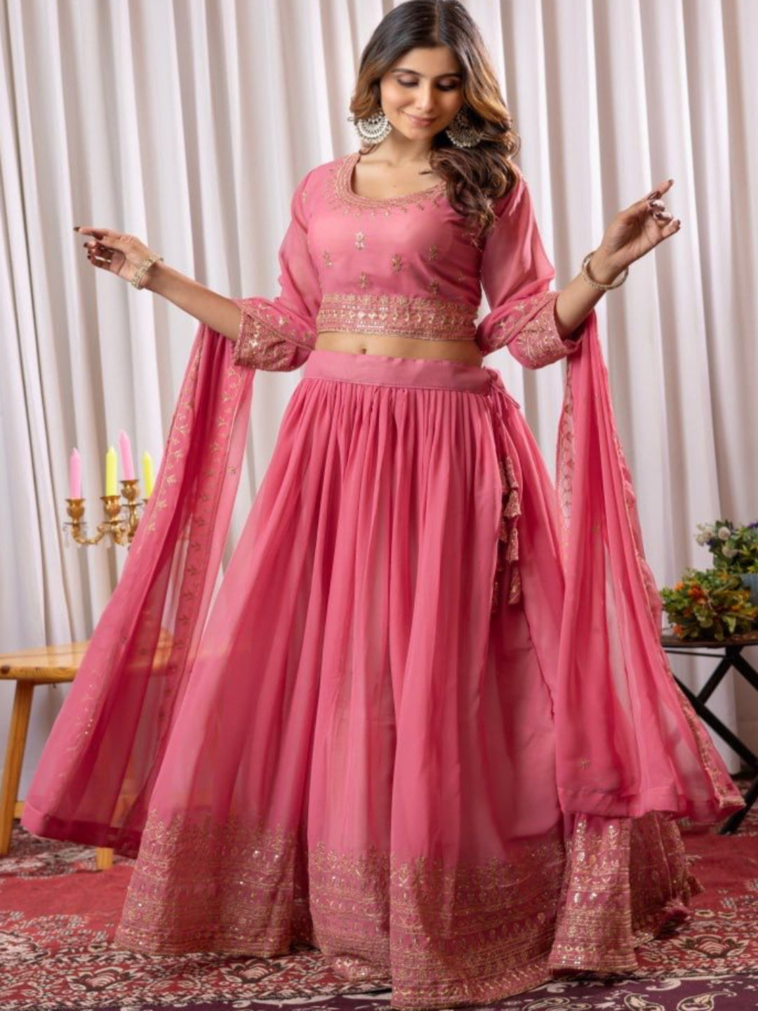 Woman in a pink traditional outfit standing in a decorated room with curtains and a chair.