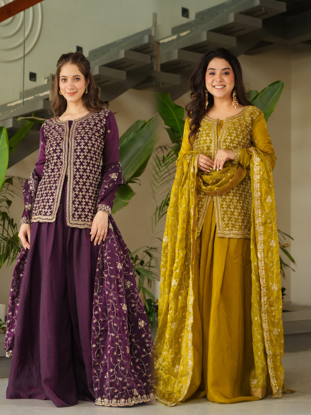 Two women in traditional outfits standing indoors with plants in the background