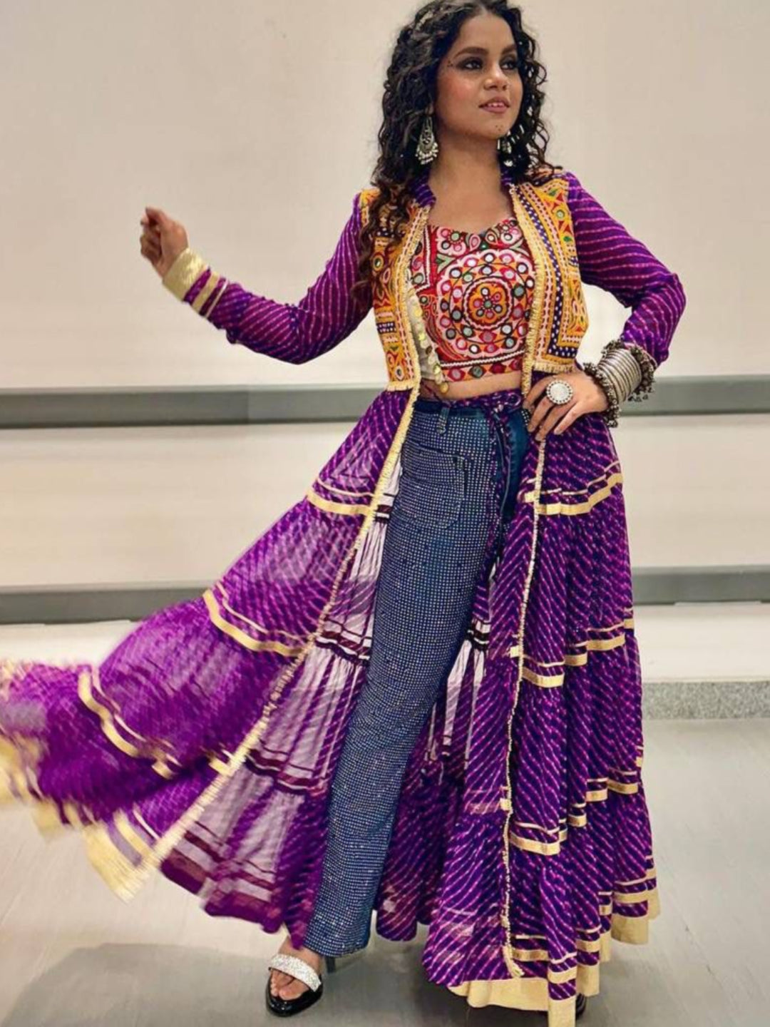 Woman in a traditional outfit with a purple and gold dress and blue jeans, standing indoors.
