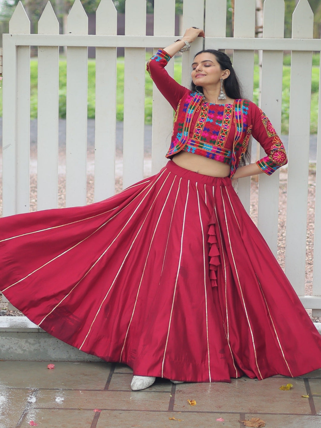 Woman in traditional outfit with red skirt and multicolored top against a white picket fence.
