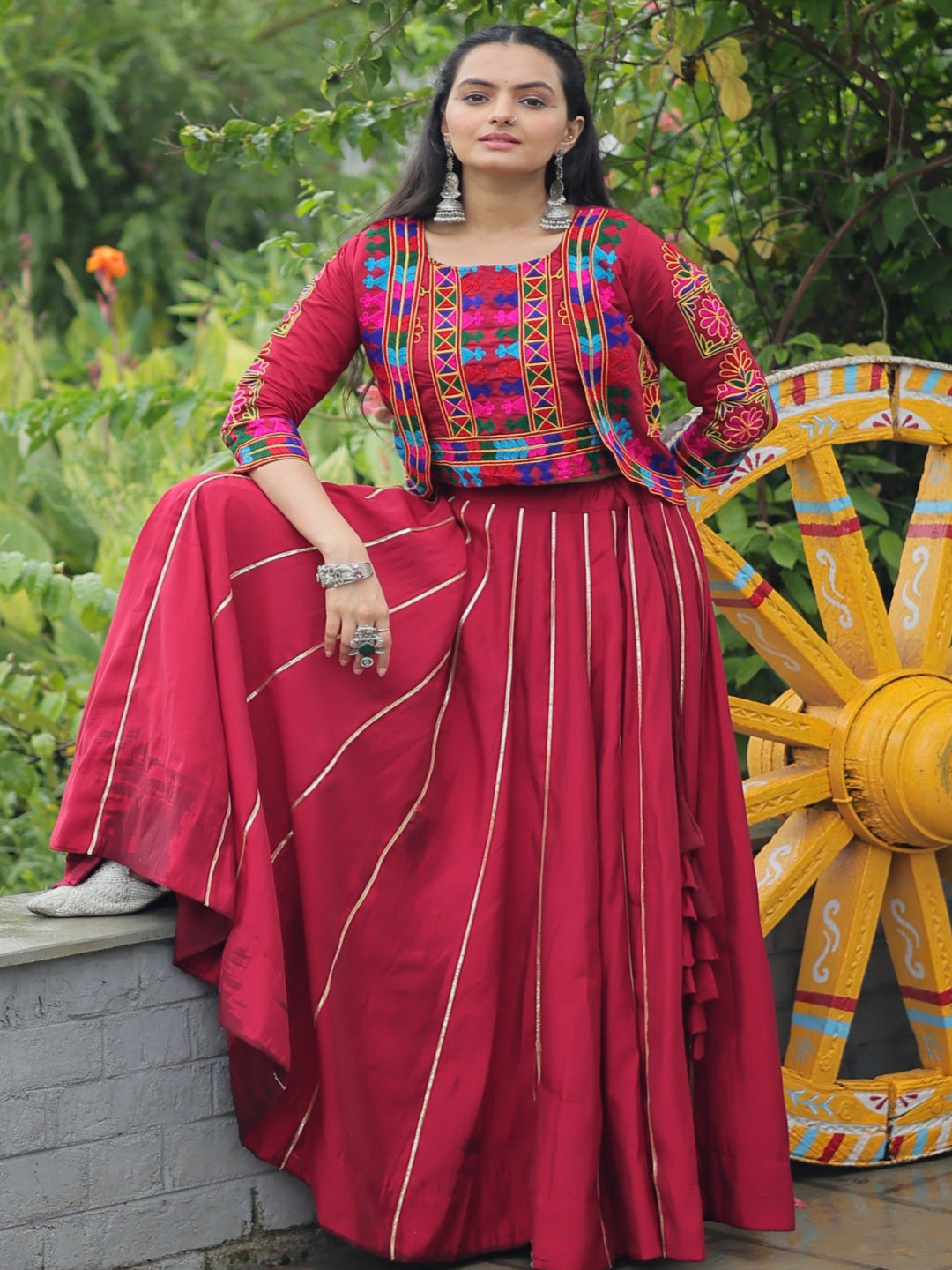 Woman in traditional outfit sitting near a colorful wheel with greenery in the background