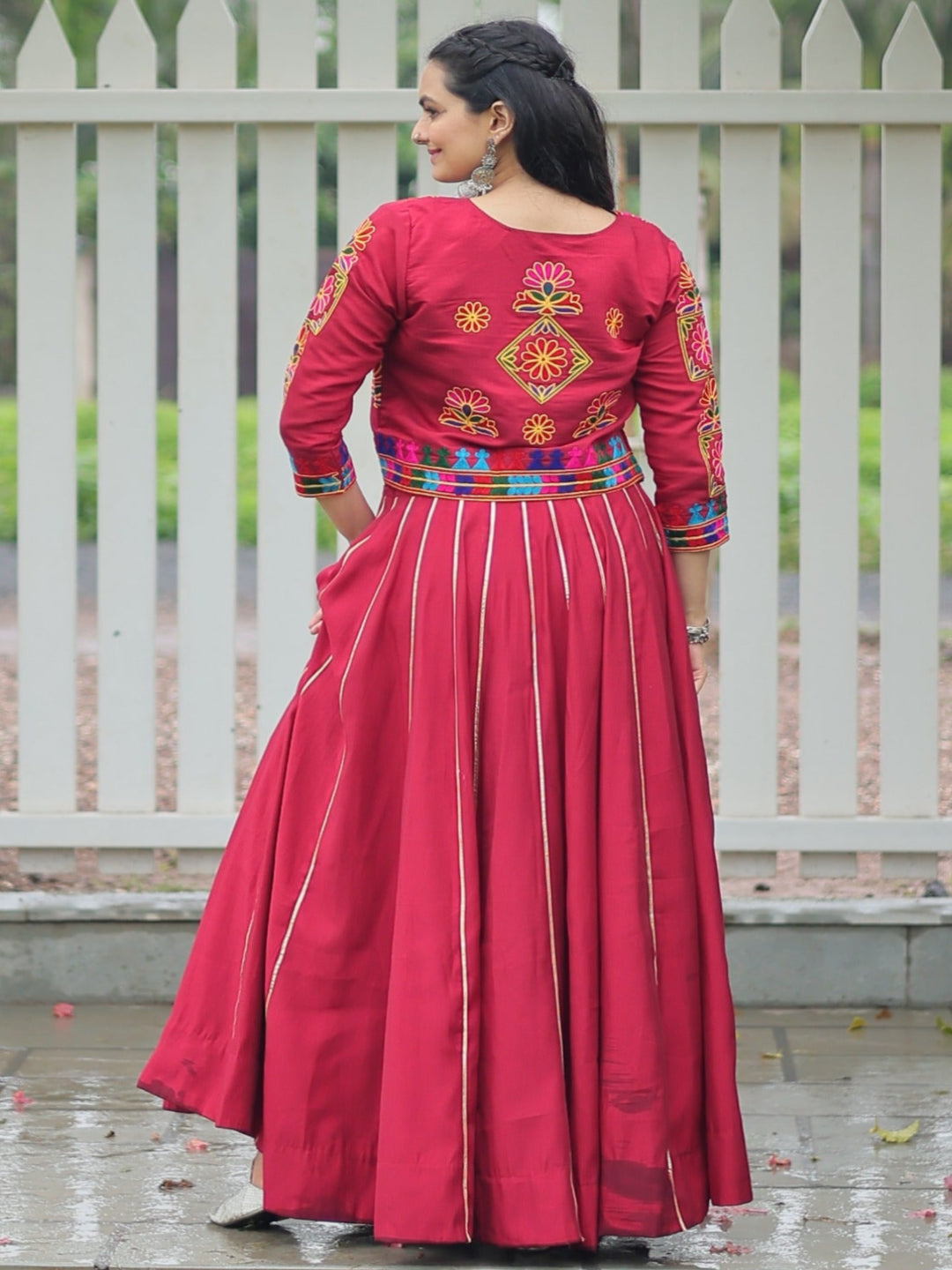 Woman in a red traditional outfit standing in front of a white picket fence.