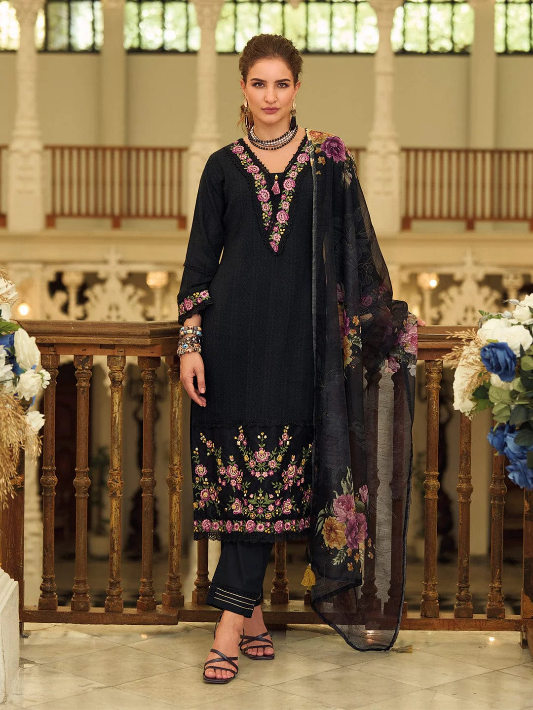 Woman wearing a black embroidered outfit with a floral dupatta in an elegant indoor setting.