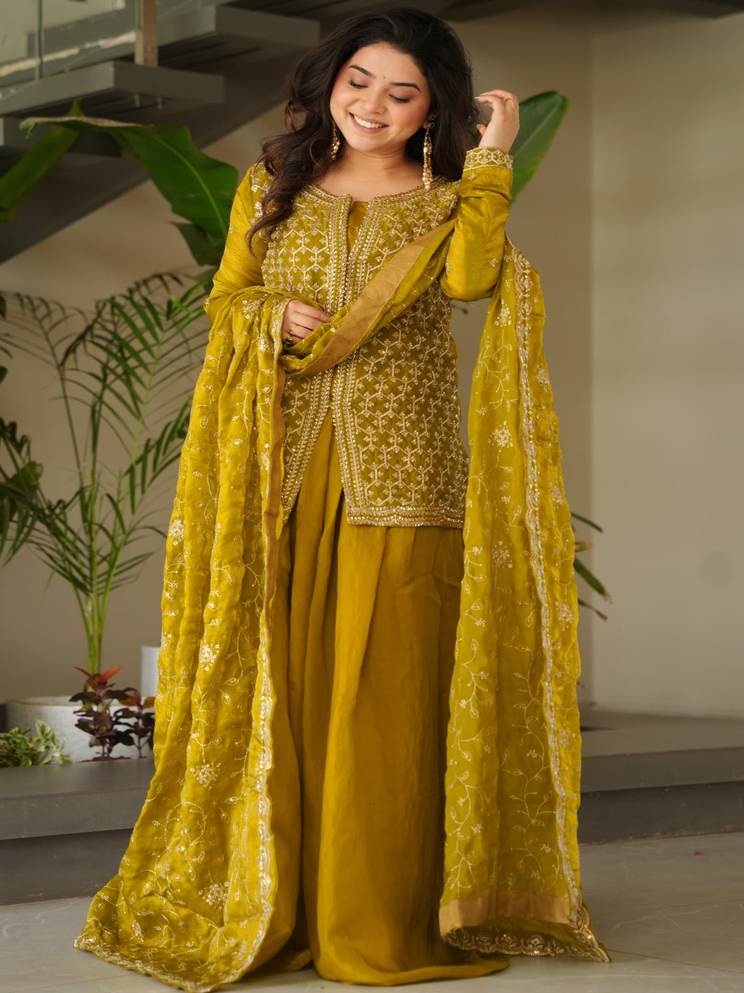 Woman in a yellow traditional outfit standing indoors with plants in the background