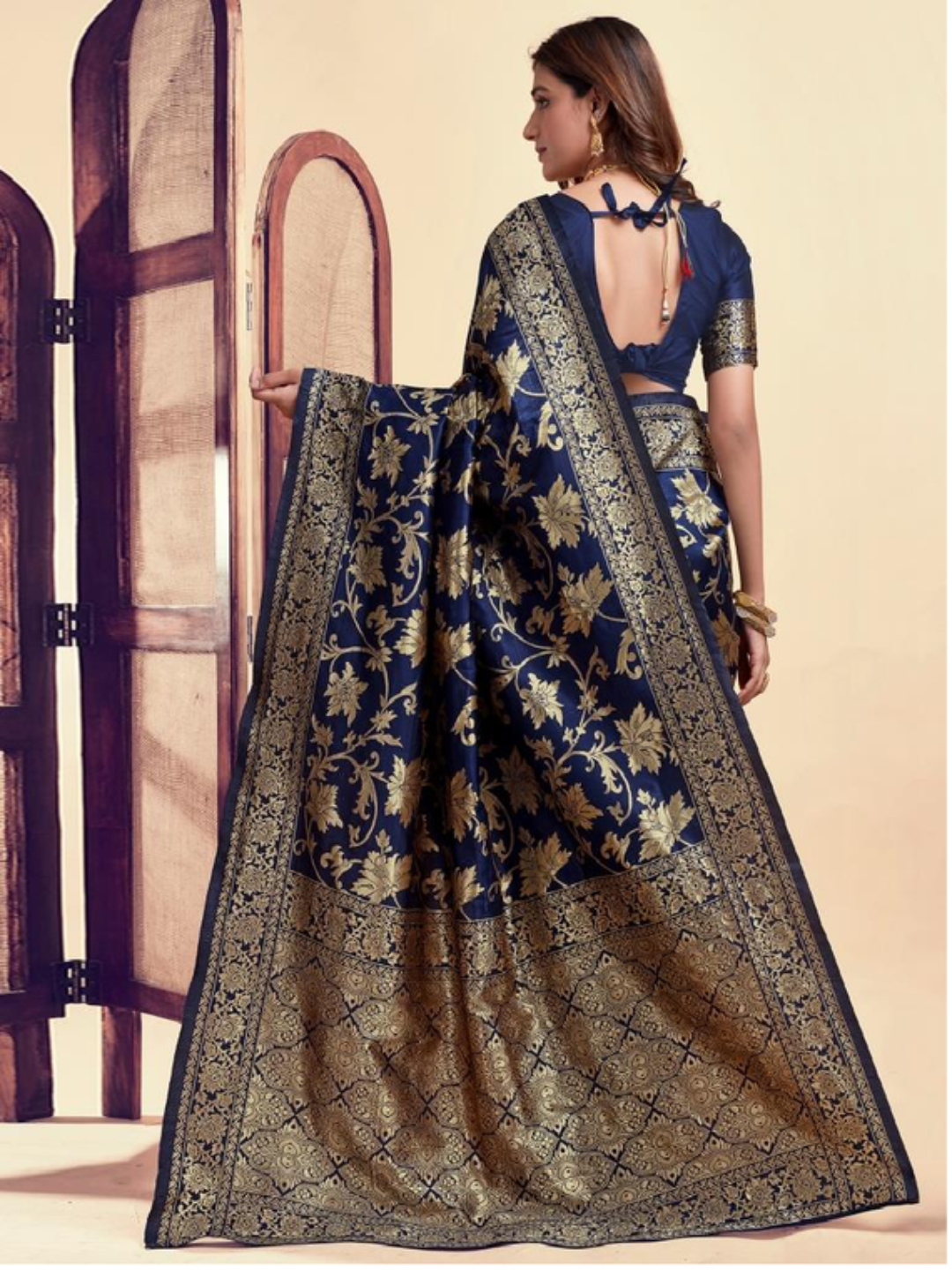 Woman wearing a blue and gold saree with a floral pattern, standing in front of a wooden door.