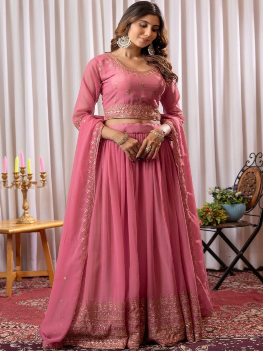 Woman in a pink traditional outfit standing in a decorated room with curtains and a table.