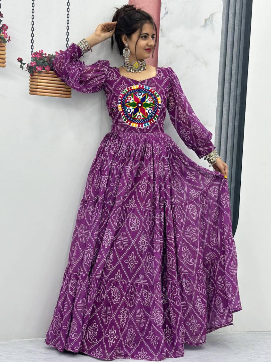 Woman wearing a purple dress with intricate patterns and a colorful center design, standing against a white wall.