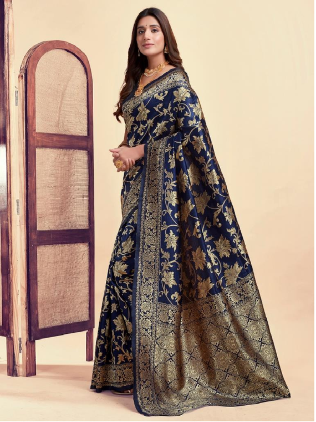 Woman wearing a blue and gold saree with a decorative pattern, standing next to a wooden mirror.