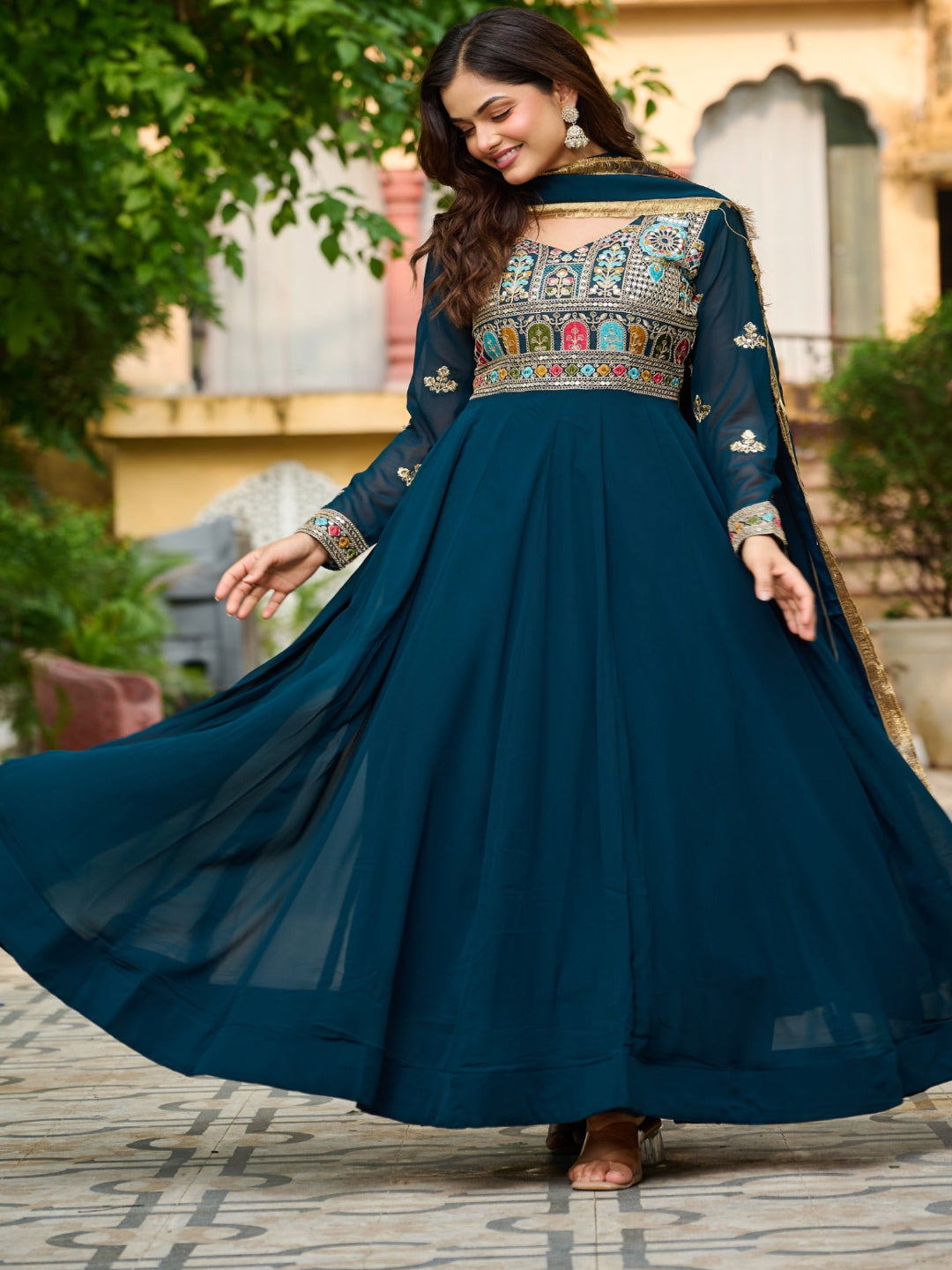 Woman in a teal traditional outfit with intricate patterns standing outdoors.