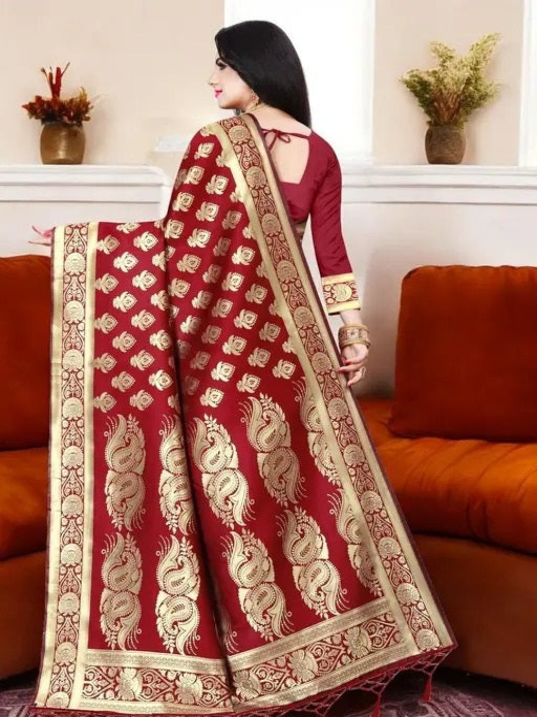 Woman wearing a red saree with gold patterns in a room with orange sofas and decorative plants.