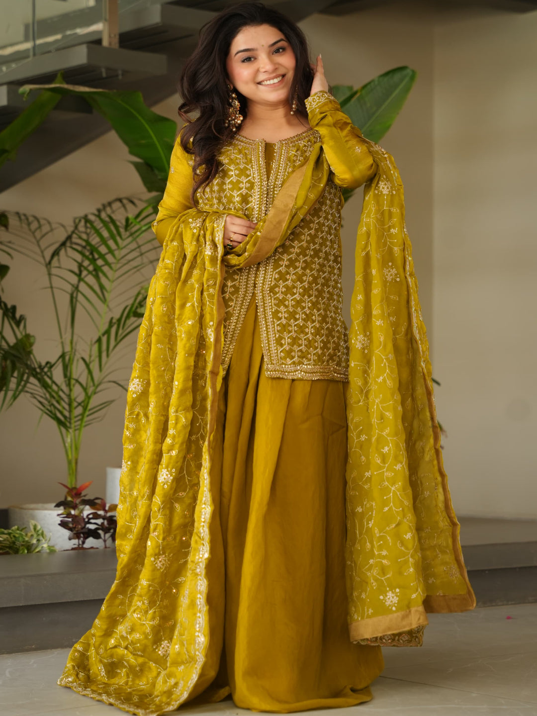 Woman in a yellow traditional outfit standing indoors with plants in the background