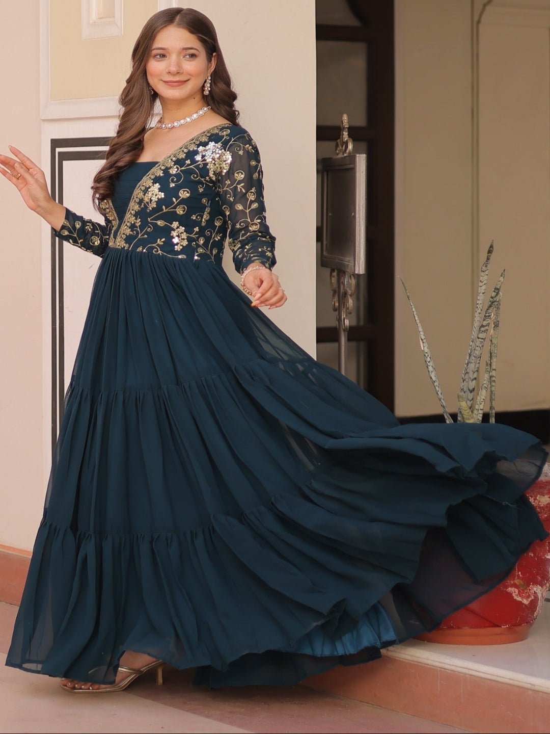 Woman in a dark blue embroidered gown standing in an ornate interior setting.