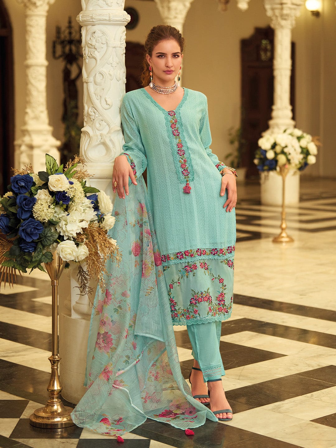 Woman in a light blue embroidered outfit standing in an elegant interior setting with floral decorations.