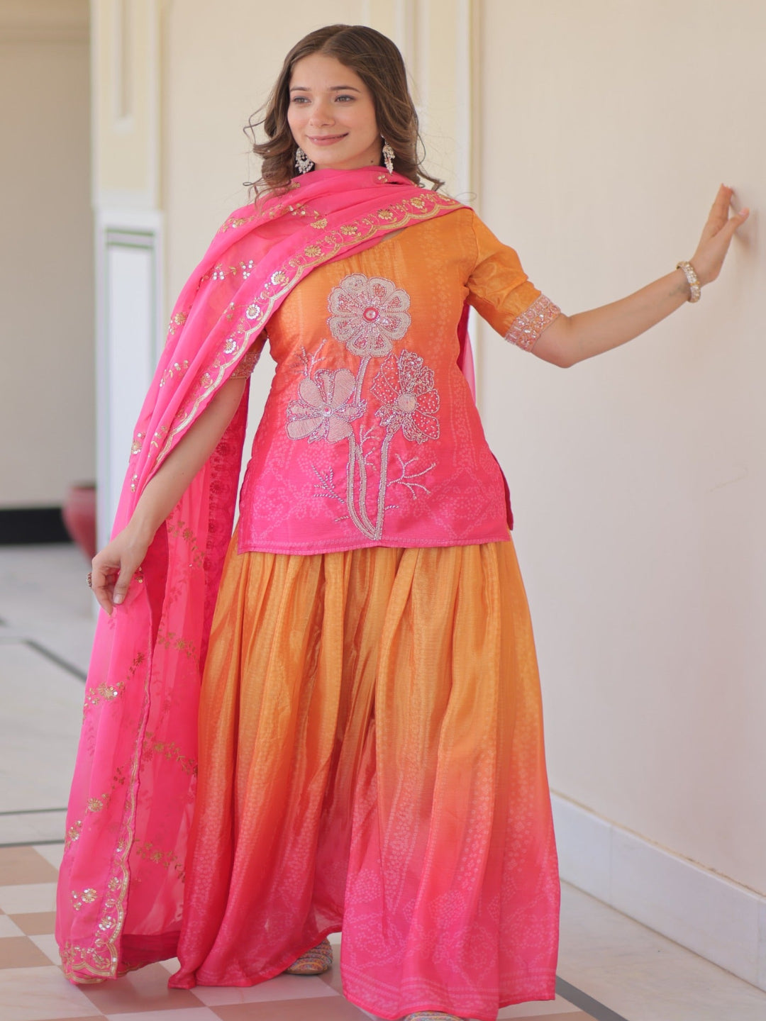 Woman wearing a pink and orange traditional outfit with a matching dupatta indoors.