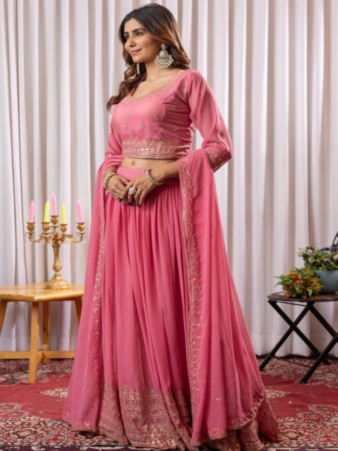 Woman in a pink traditional outfit standing in a decorated room with candles and a plant.