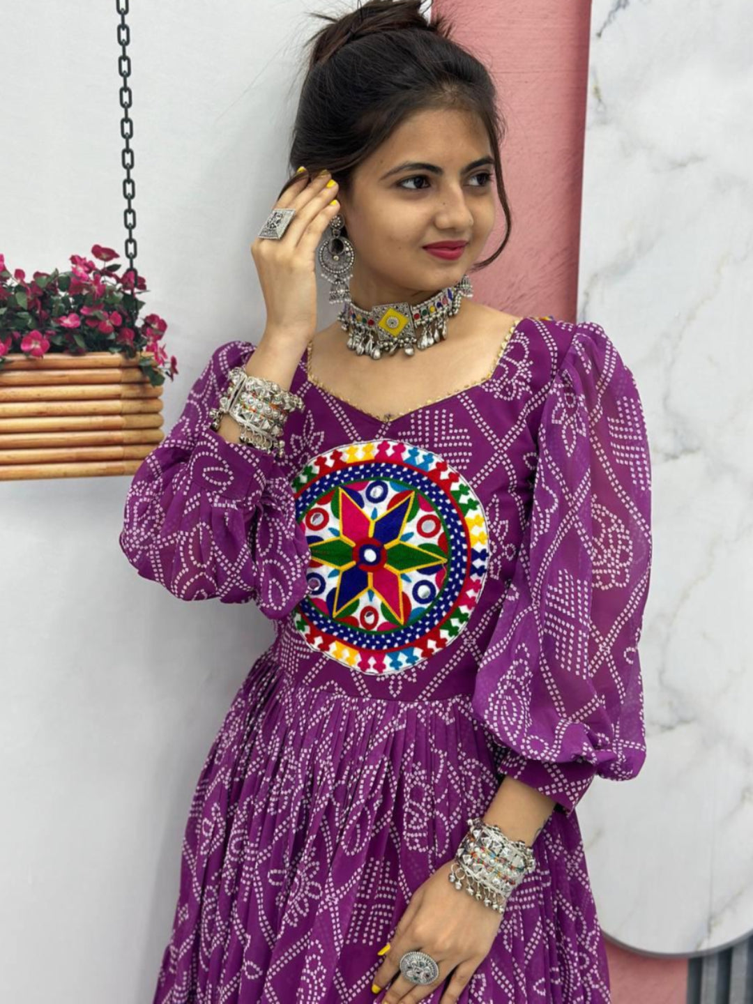 Woman wearing a purple dress with colorful design, standing against a white wall with pink and wooden elements.