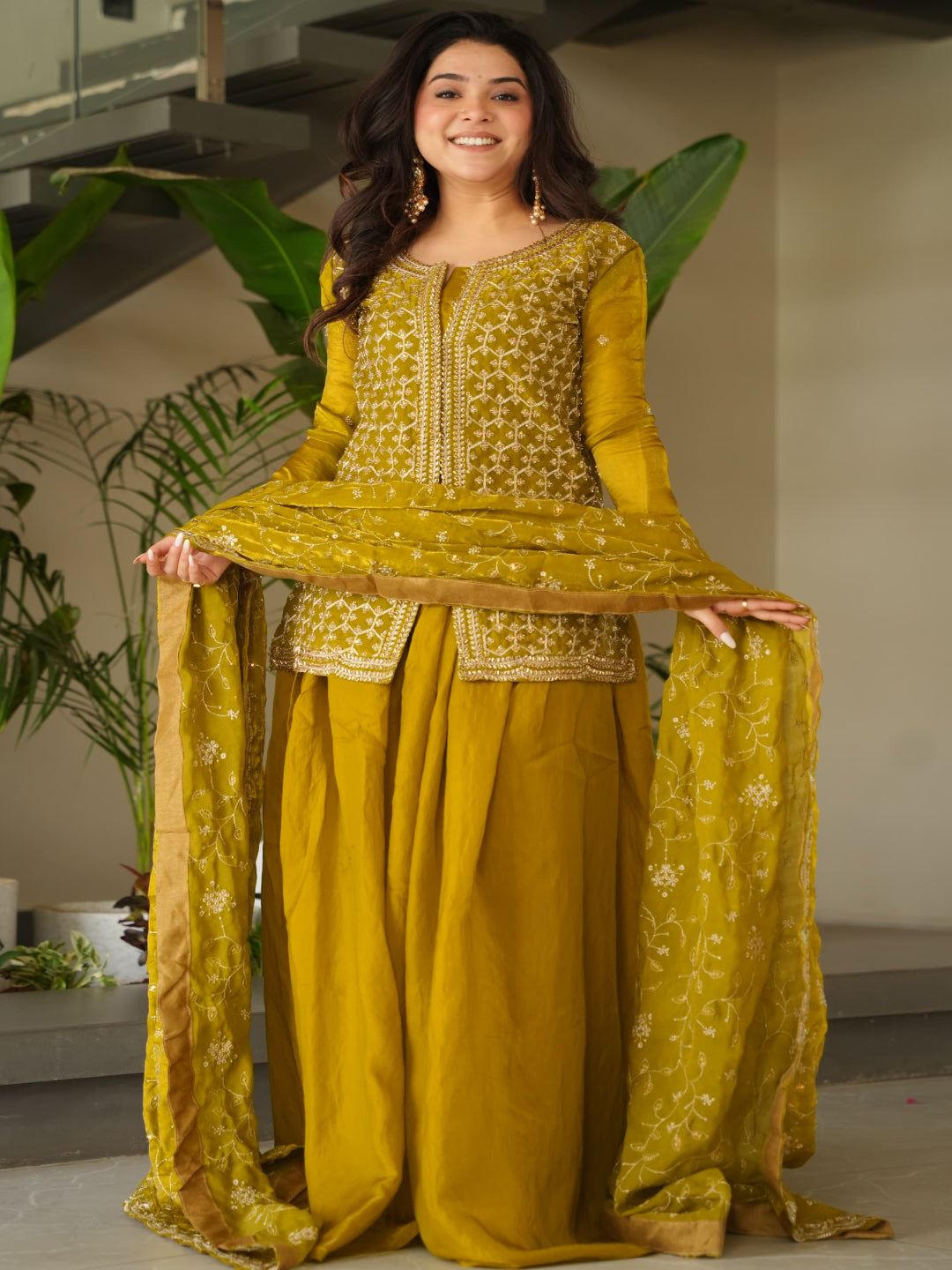 Woman in a yellow traditional outfit standing indoors with plants in the background