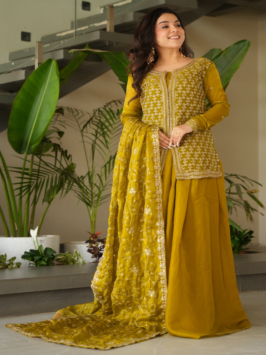 Woman in a yellow traditional outfit standing indoors with plants in the background