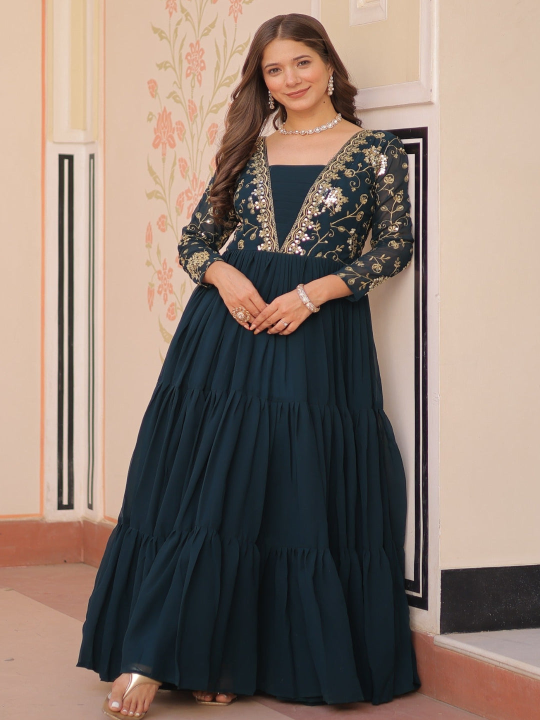 Woman in a dark blue embroidered dress standing indoors against a decorative wall.