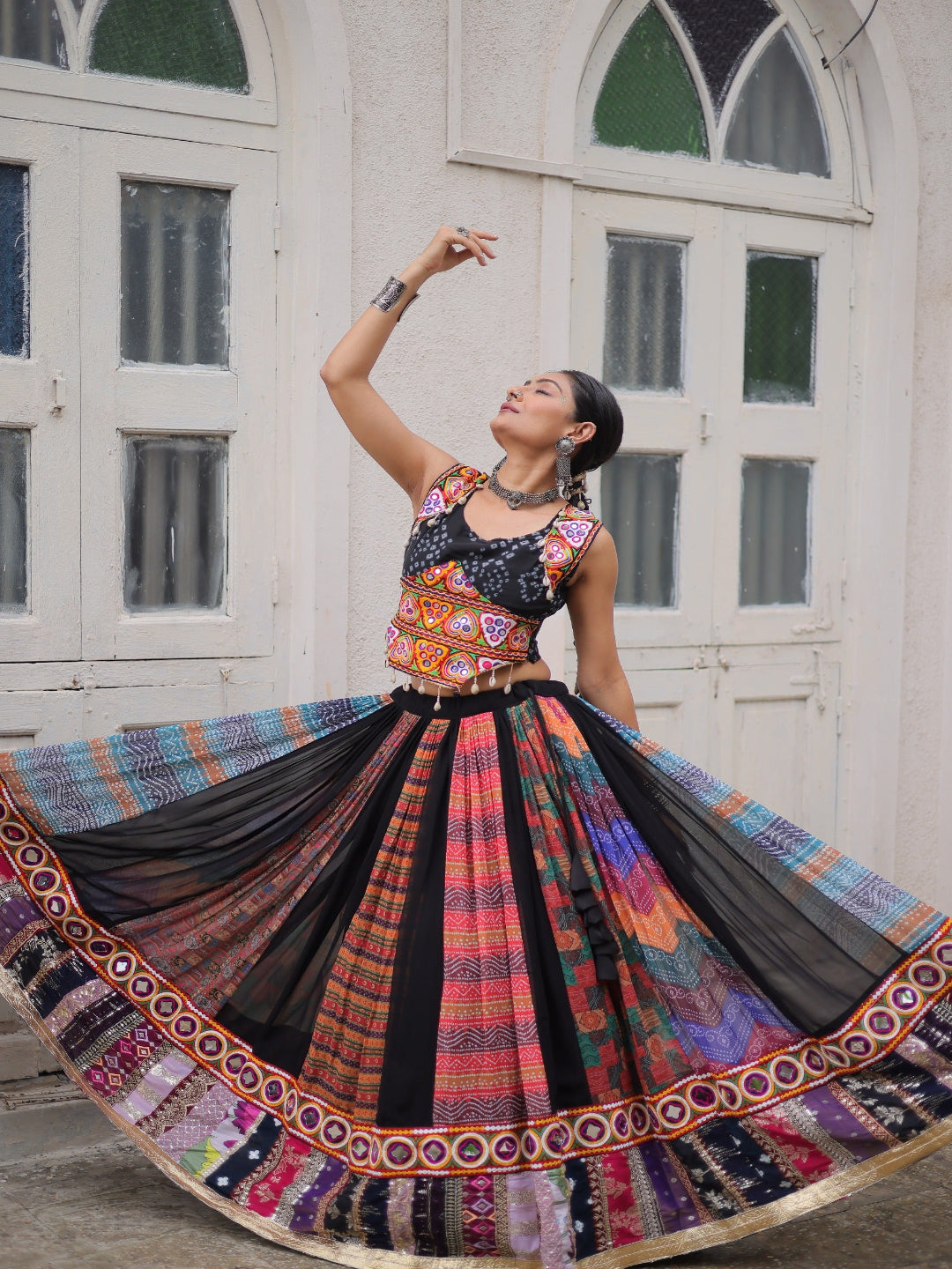 Woman in a colorful traditional outfit with a long skirt, standing in front of a white building with arched windows.