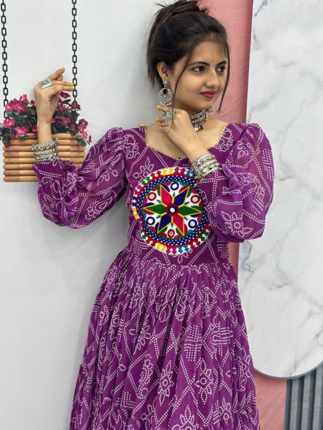 Woman wearing a purple dress with a colorful floral design, standing against a white wall.