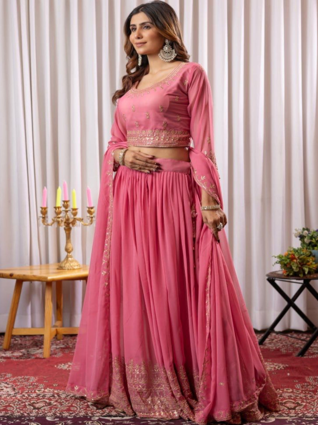 Woman in a pink traditional outfit standing in a decorated room with a white curtain background.