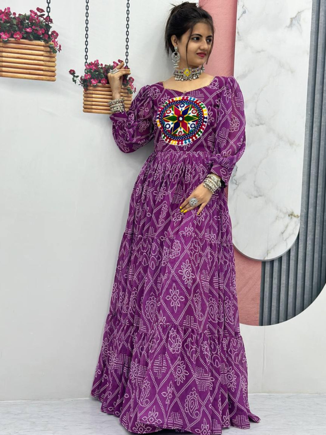 Woman wearing a purple dress with a colorful pattern, standing against a white wall.