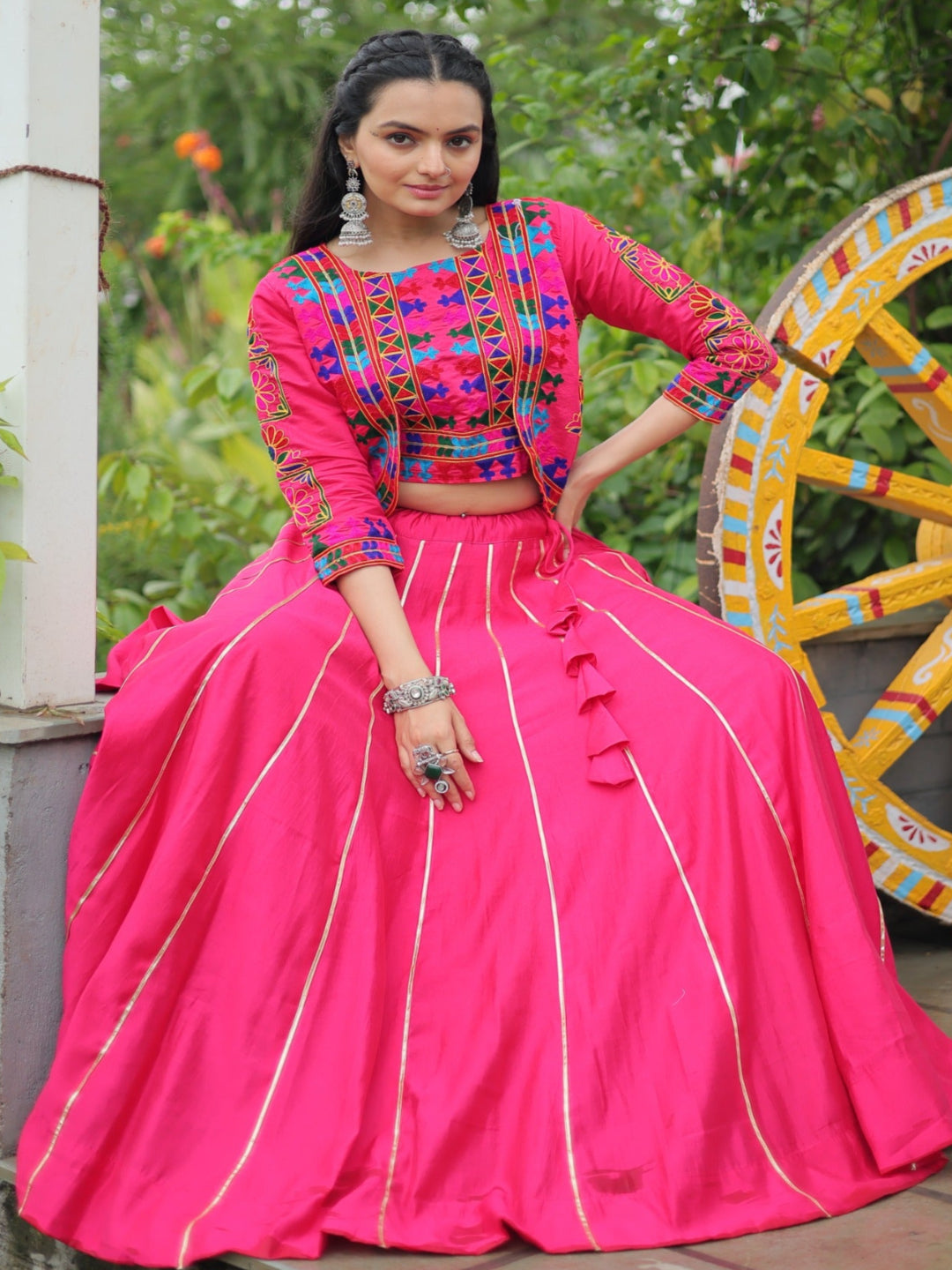 Woman in a bright pink traditional outfit with colorful blouse, sitting next to a large wheel with decorative patterns.
