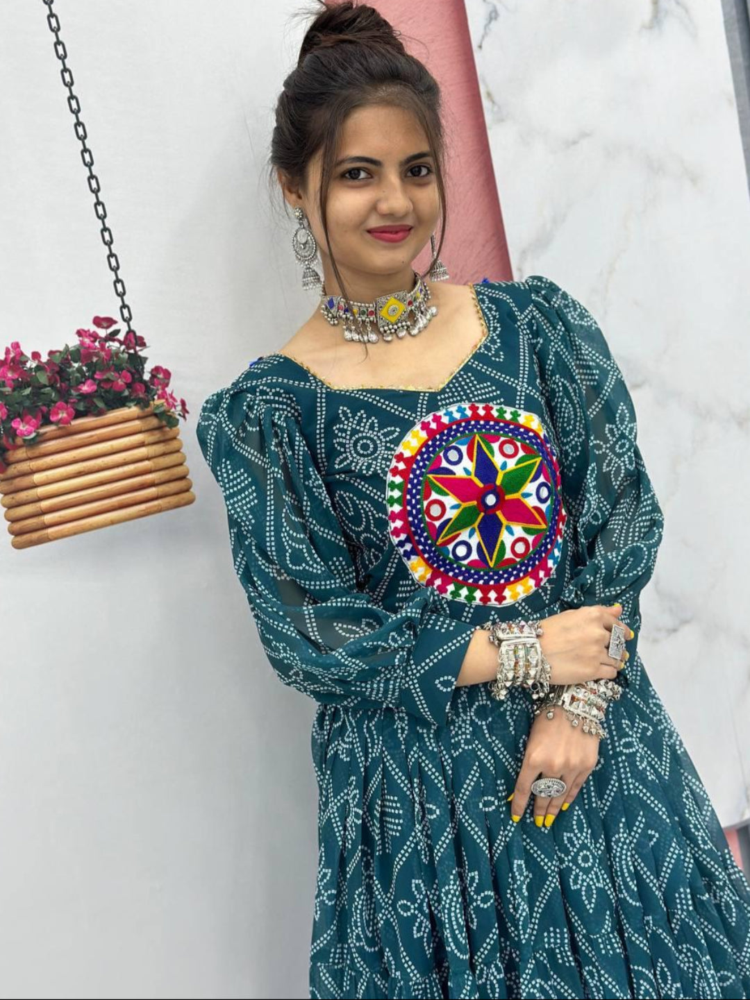 Woman wearing a green traditional outfit with a colorful embroidered design, standing against a white wall with a hanging plant.