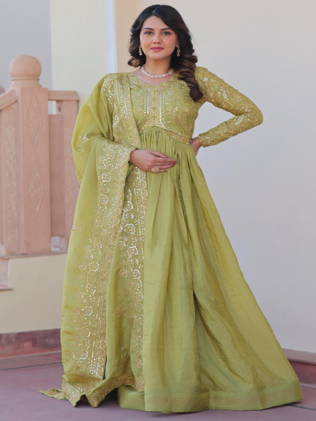 Woman in a green traditional outfit standing indoors with a staircase in the background