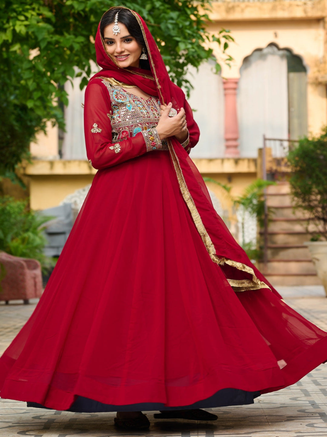 Woman in a red traditional outfit standing outdoors with greenery and architectural elements in the background