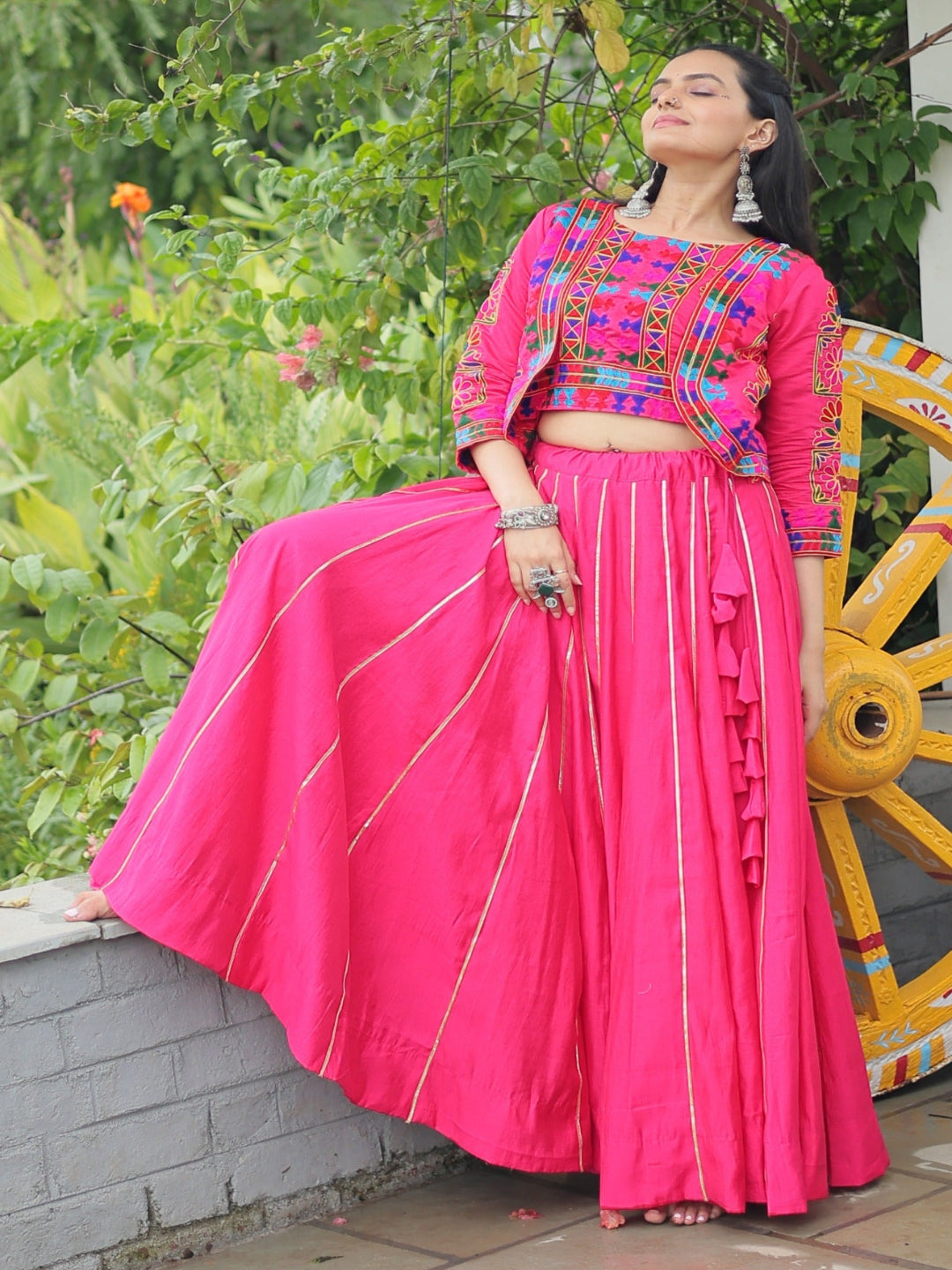 Woman in a colorful traditional outfit sitting near a decorative wheel with greenery in the background