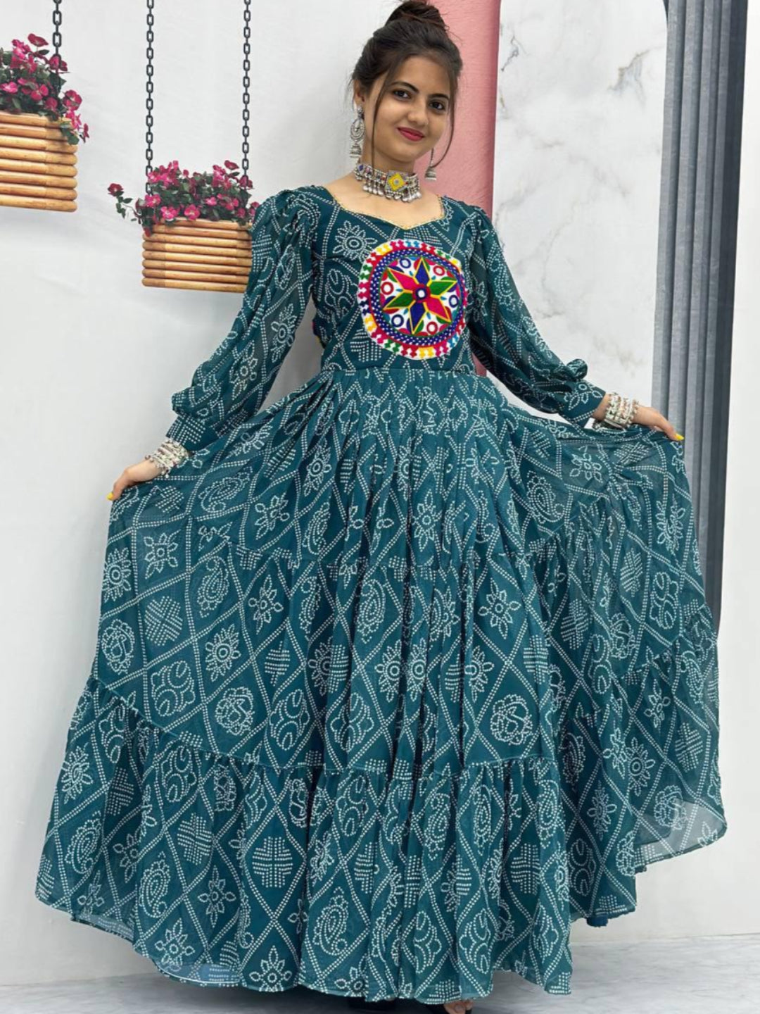 Woman wearing a teal patterned dress with a colorful embroidered top, standing indoors.
